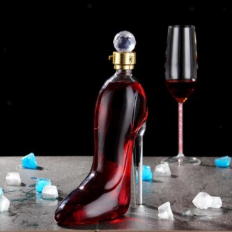 Elegant wine bottle shaped like a high-heeled shoe, filled with vibrant red liquid, placed on a table with scattered ice cubes and a champagne flute in the background